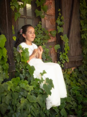 una niña preciosa rodeada de verde el dia de su primera comunión