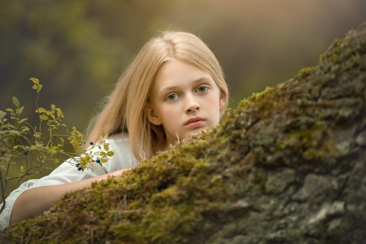 foto de niña rubia con ojos azules en vestido blanco de primera comunión apoyada en un árbol