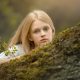 foto de niña rubia con ojos azules en vestido blanco de primera comunión apoyada en un árbol